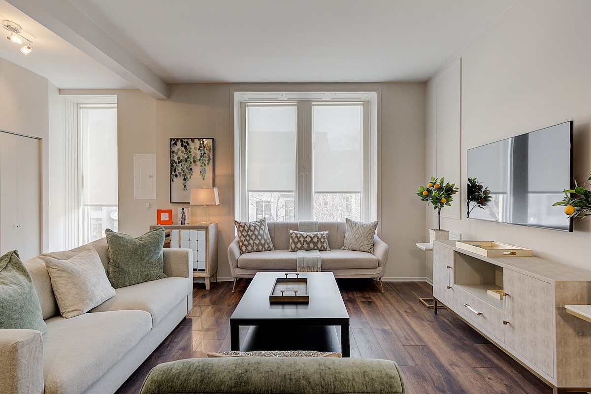 A spacious living area is highlighted by large windows that allow natural light to fill the room. Two comfortable sofas are arranged around a central coffee table. A flat-screen television is mounted on the wall opposite the seating, while a side table holds a decorative lamp and art piece.