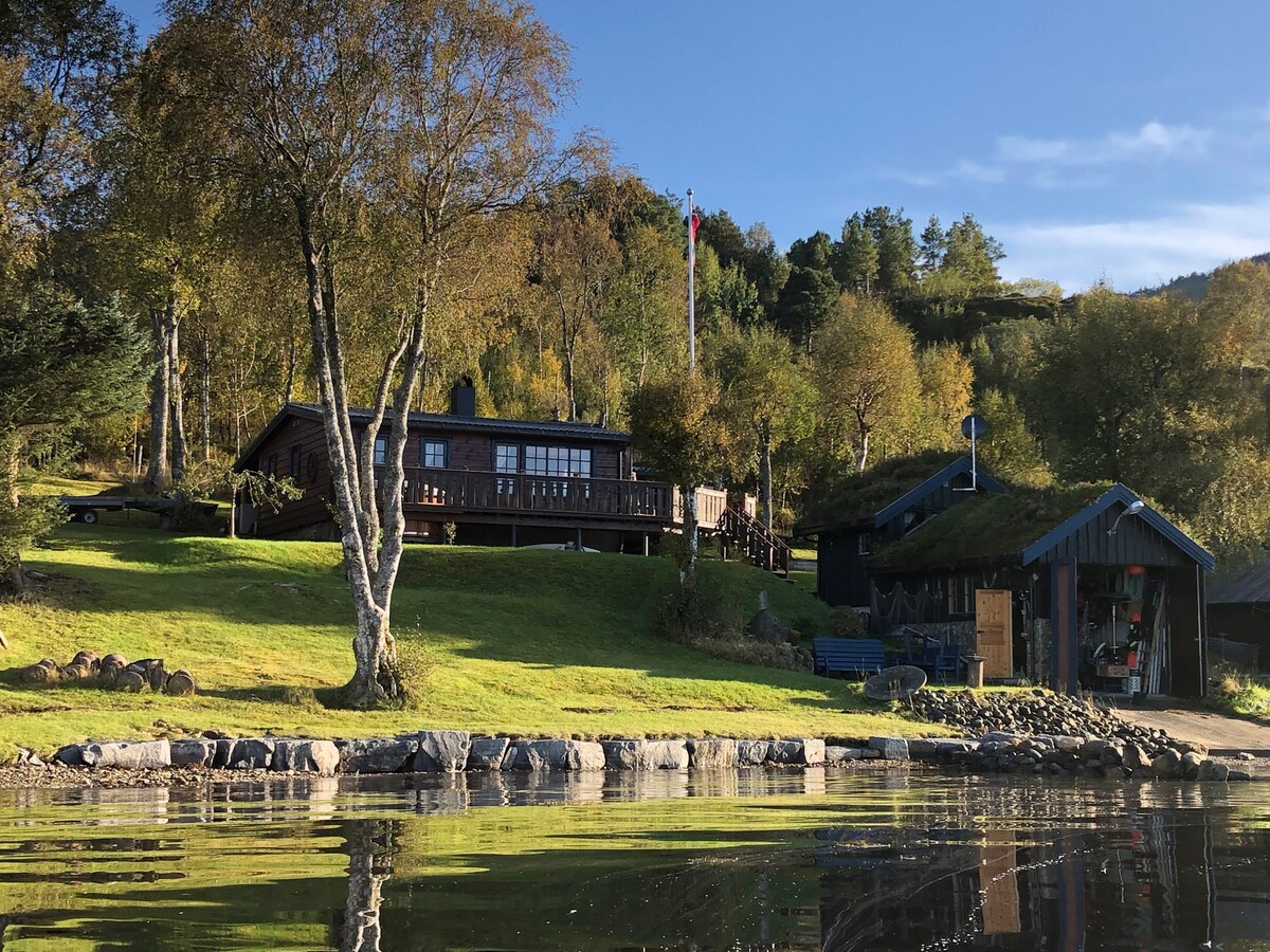 A wooden cabin is positioned near the water, surrounded by greenery and trees. A spacious terrace extends from the cabin, offering outdoor seating. The calm water reflects the surrounding landscape and the clear blue sky, creating a serene environment.
