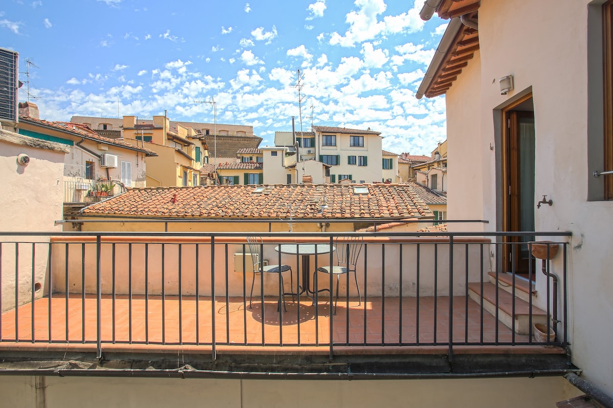 A small terrace is depicted with a view of the rooftops of Florence, featuring a round table and two chairs. The red-tiled rooftops and blue sky with scattered clouds create a welcoming outdoor space.