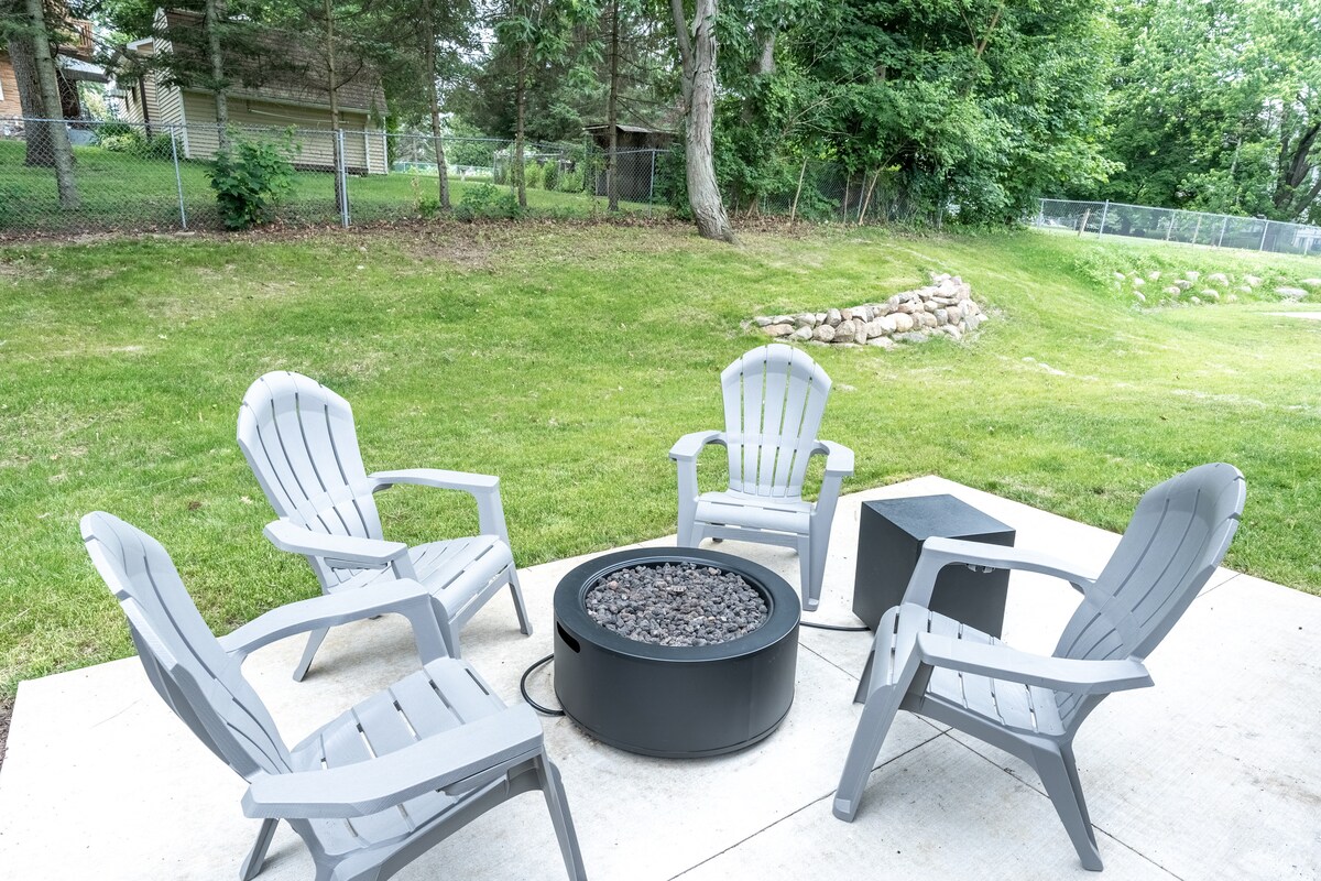 A patio area features five light-colored Adirondack chairs arranged in a circle around a modern gas-powered fire pit. A grassy expanse extends beyond the seating, framed by trees and a low stone wall in the background.