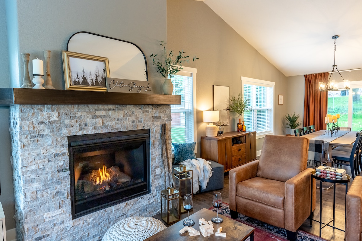 A modern living room features a stone fireplace with a lit flame, providing warmth to the cozy space. A comfortable brown armchair is positioned nearby, complemented by a wooden side table. Natural light filters through the windows, enhancing the welcoming atmosphere.