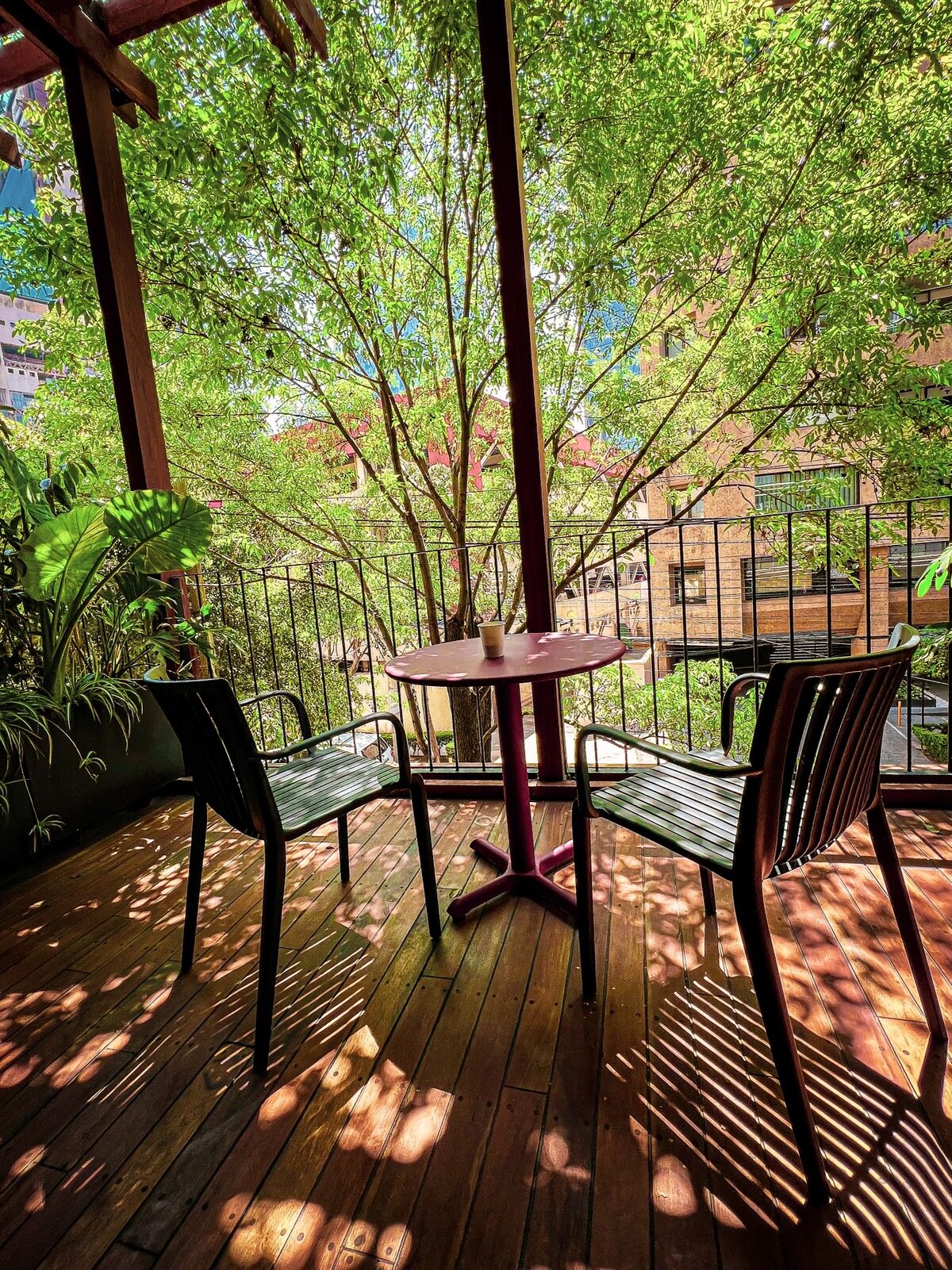 A small terrace is surrounded by lush greenery, featuring two black chairs and a round wooden table. Gentle sunlight filters through the leaves, creating a play of shadows on the wooden floor. The view beyond includes a vibrant cityscape.