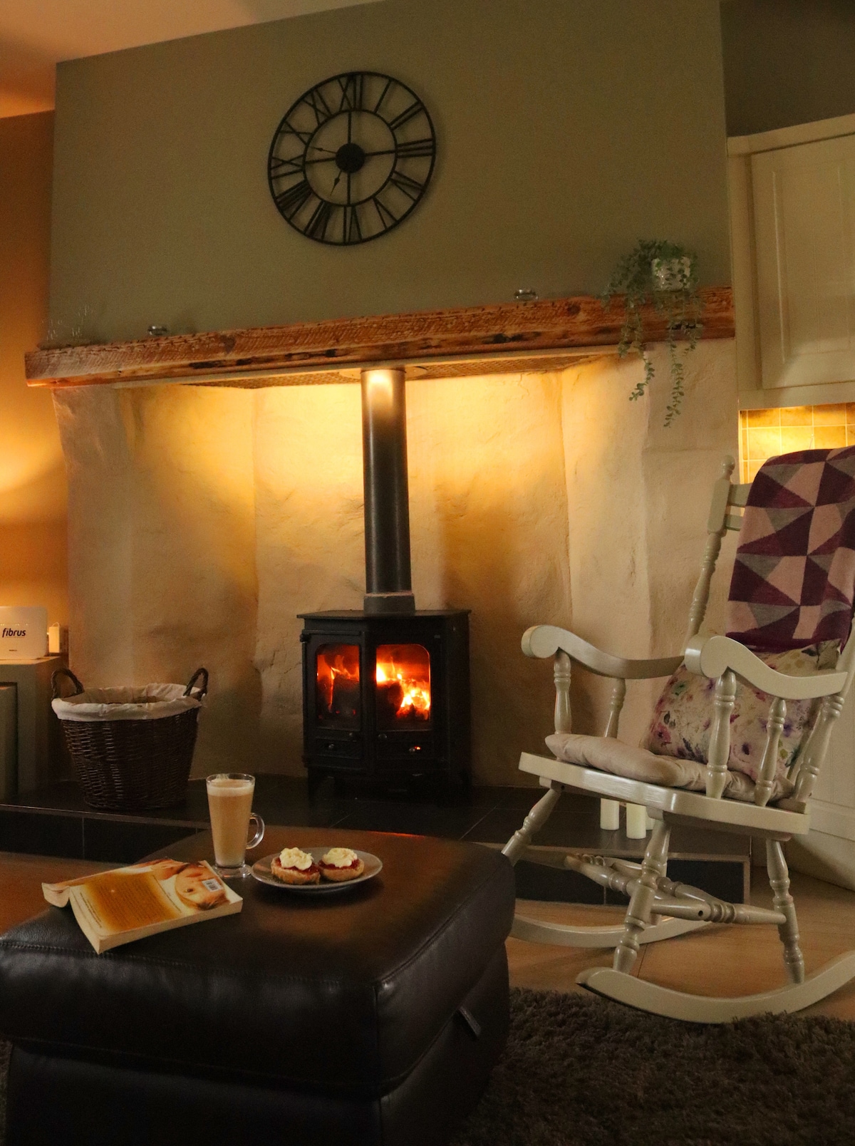 A cozy living area features a wood-burning stove set against a textured wall. A rustic clock adorns the mantel above. Nearby, a comfortable rocking chair complements a small ottoman with a tray of pastries and a beverage, creating an inviting space for relaxation.