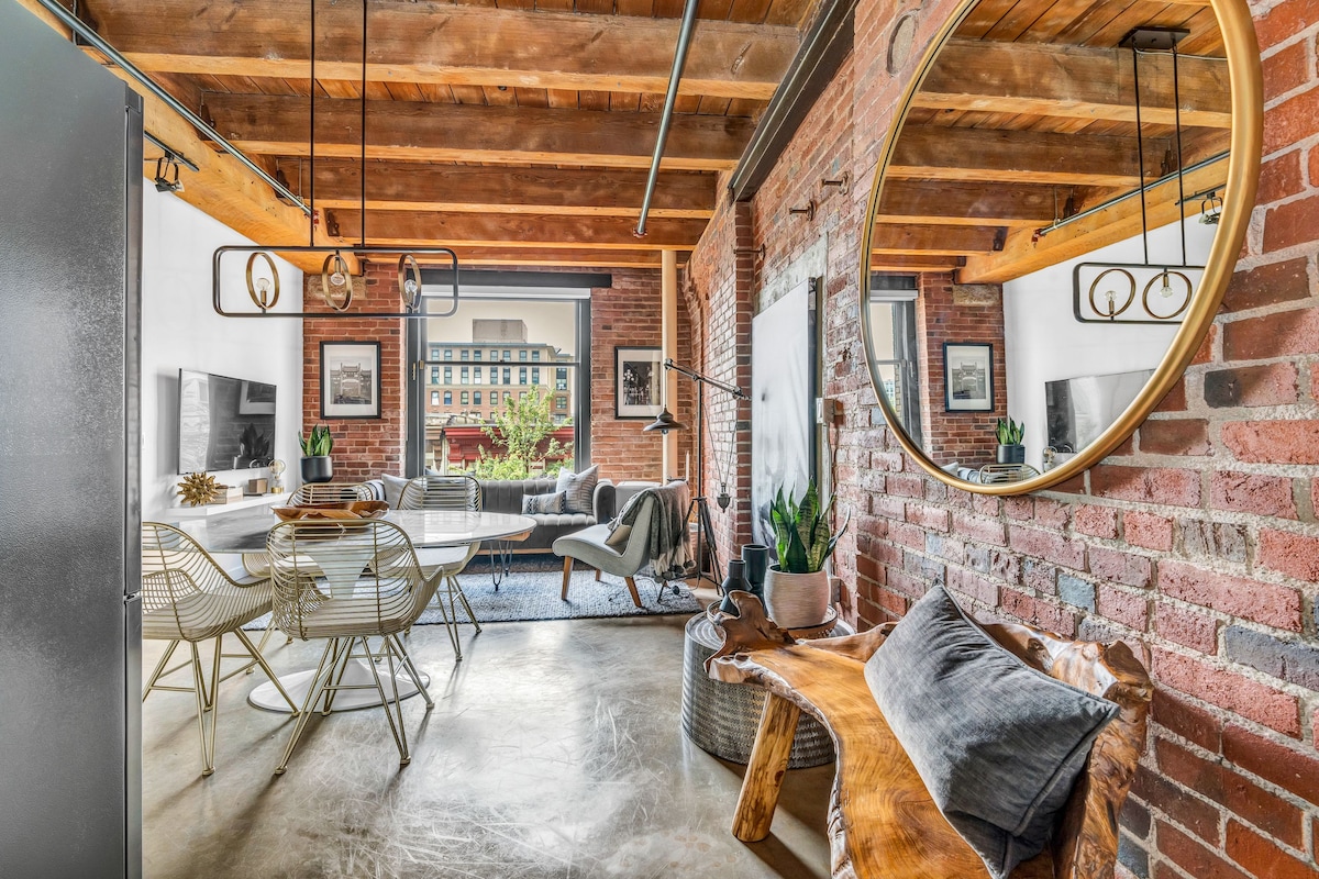 The open-concept living area features warm brick walls and exposed wooden beams, creating an industrial ambiance. A large circular mirror reflects light, while comfortable seating is arranged around a stylish rug. Lush greenery adds life to the space, complemented by natural light from large windows.