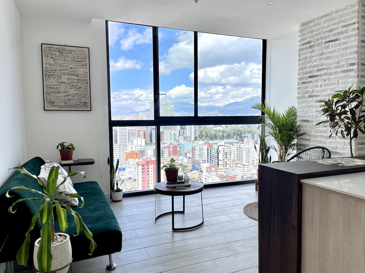 A modern living space is presented with large floor-to-ceiling windows offering a panoramic view of the city. A green sofa complements decorative plants, while light-colored walls enhance the bright ambiance. The flooring features light wood tones, and a geometric rug adds subtle texture.