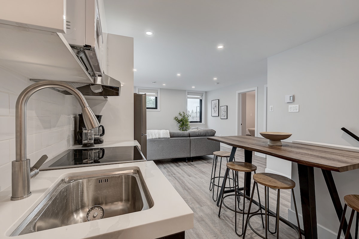 A modern kitchen area is accented by sleek cabinetry and stainless steel appliances. An island with bar stools offers additional seating. In the background, a living area can be seen, featuring a comfortable sofa and decorative wall art.
