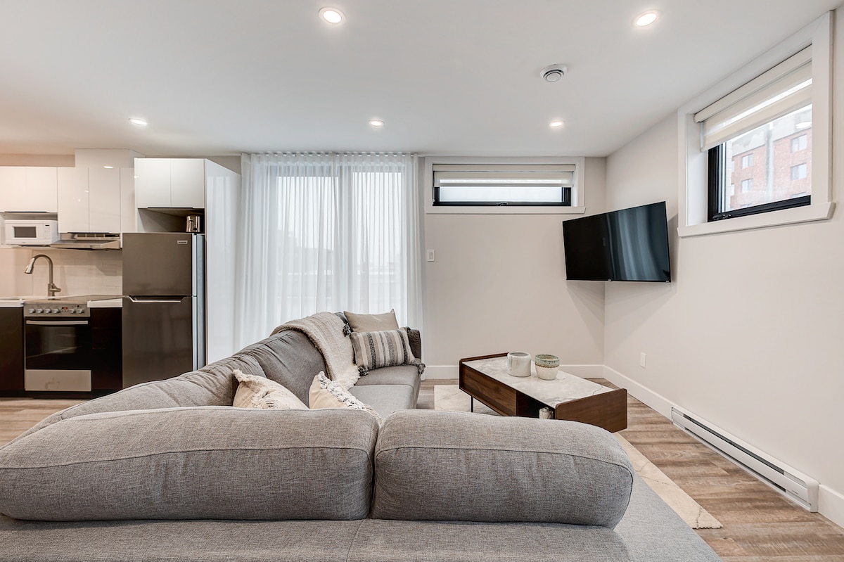 A comfortable living area is presented with a spacious sectional sofa, a wooden coffee table, and a mounted flat-screen television. Natural light filters through sheer curtains, illuminating the modern kitchen appliances and cabinetry in the background.