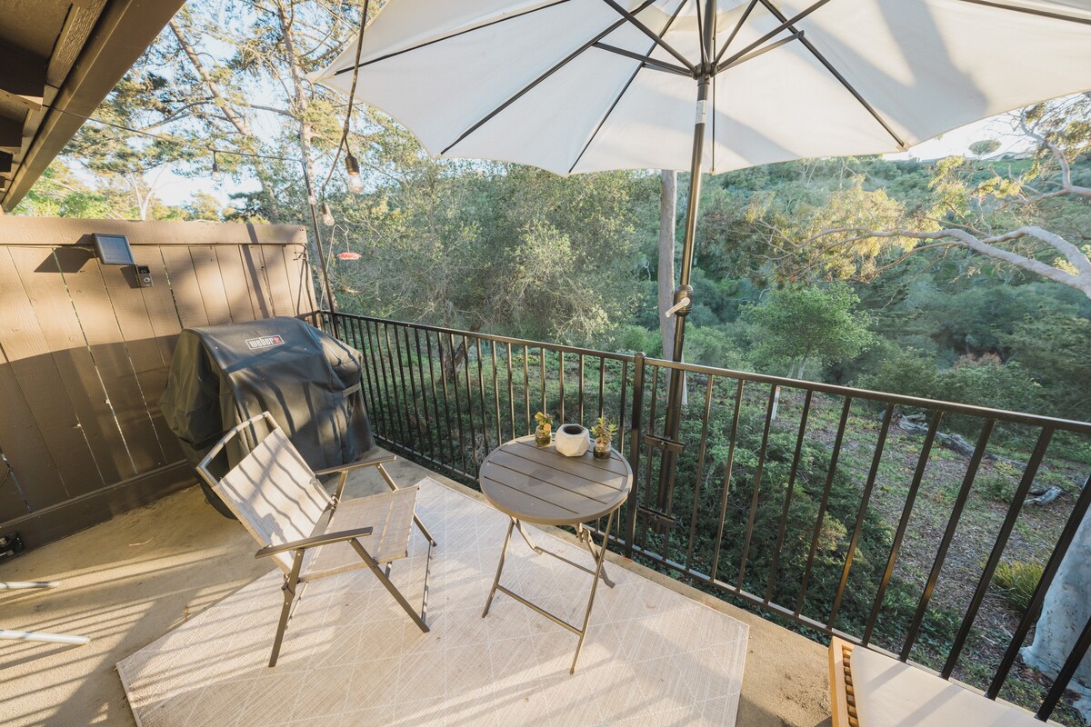 A private balcony space is presented with a small round table and a folding chair under a large umbrella. A grill is set against the wall, surrounded by lush greenery that provides a natural backdrop.