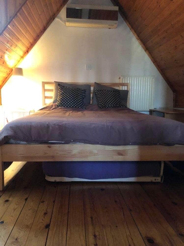 A cozy loft bedroom features a bed with a wooden frame, topped with a dark lavender blanket. Two decorative pillows are placed on the bed. The angled wooden walls create a warm atmosphere, while ambient light is provided by a lamp beside the bed.