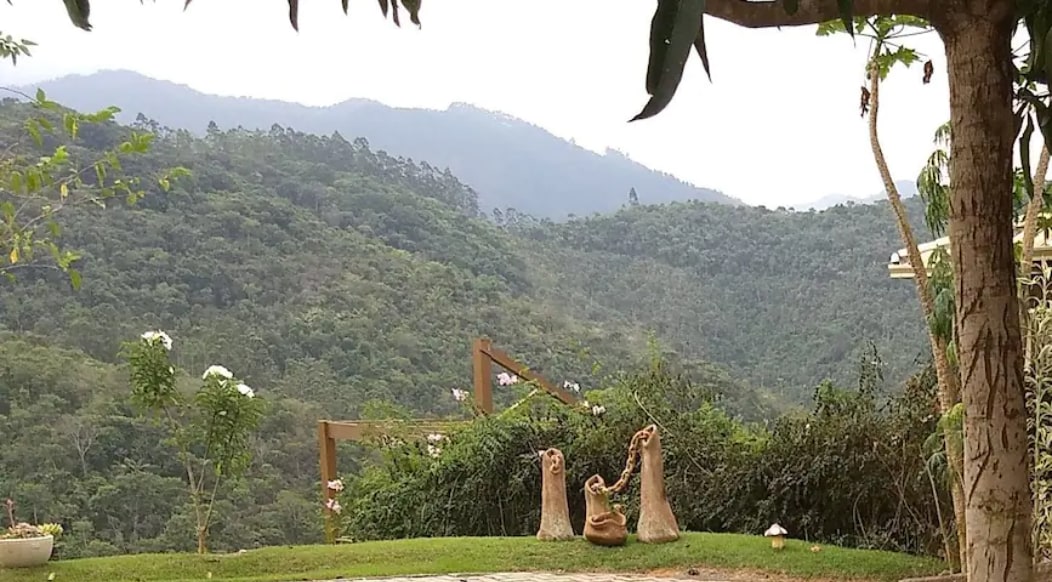 A scenic view of rolling green mountains is captured in the distance, framed by lush trees and vibrant foliage. The foreground features artistic sculptures, adding an element of charm to the natural surroundings. Soft clouds are visible in the overcast sky, creating a serene atmosphere.