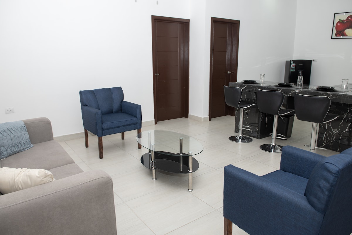 A modern living area features a light-colored sofa and two blue accent chairs surrounding a glass coffee table. A sleek black bar with stools is visible in the background, along with a doorway leading to additional rooms.