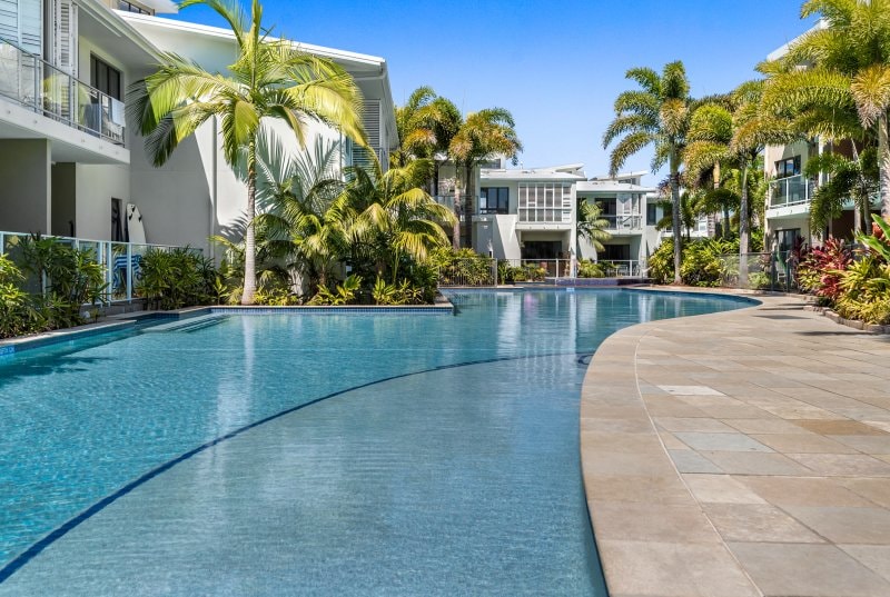 A lagoon-style swimming pool curves gracefully through lush landscaping that includes palm trees and tropical plants. Surrounding the pool, modern villas are partially visible, creating a serene and inviting atmosphere under clear blue skies.