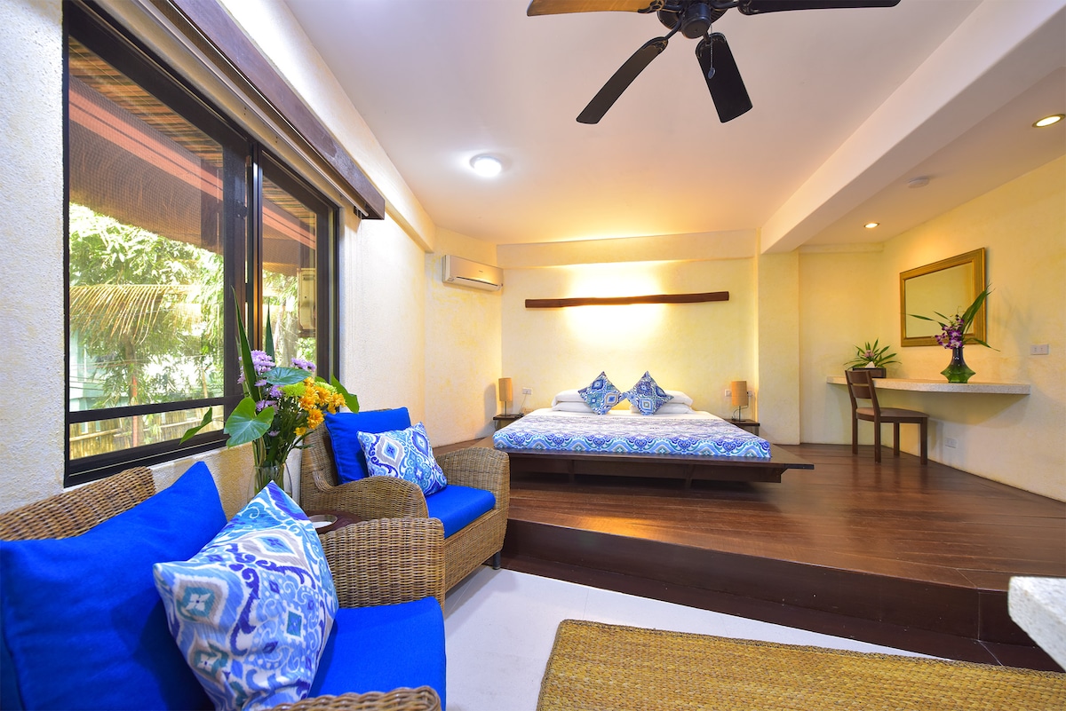 A bright sleeping area features a spacious bed adorned with blue and white decorative pillows. Comfortable seating with blue cushions is positioned near large windows, allowing natural light to fill the room. A small table and plants are visible, enhancing the inviting atmosphere.