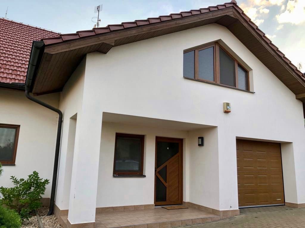 The exterior of the house features a modern architectural design with a tiled roof and a light-colored facade. A welcoming entrance is marked by a decorative door and a small porch, while a garage is visible on the right side.