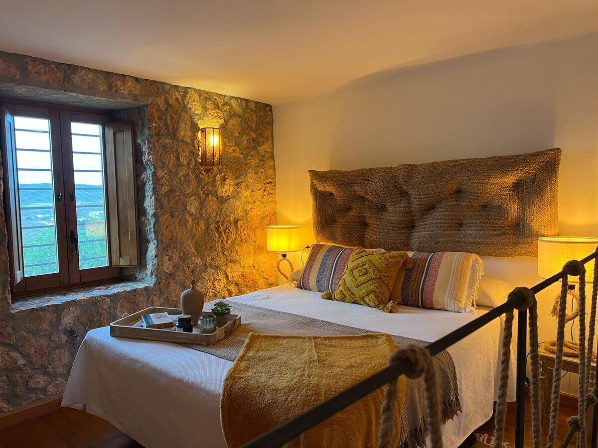 A serene bedroom features a spacious bed adorned with a textured throw and decorative pillows. Two lamps provide soft illumination beside the bed. A stone wall complements the rustic atmosphere, and a wooden window allows natural light to fill the room, revealing views of the surrounding landscape.