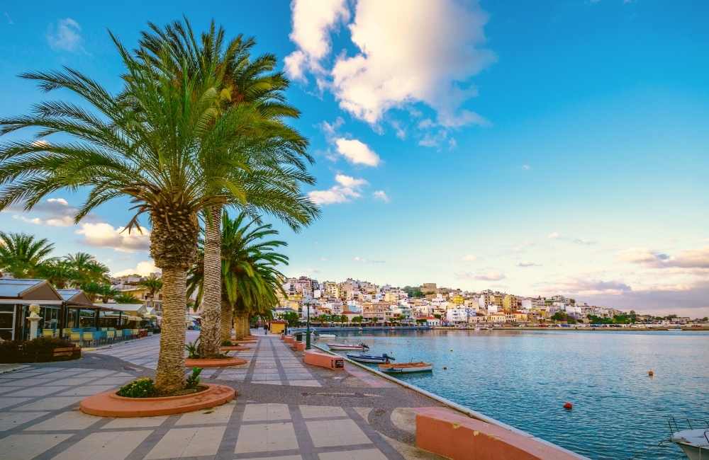 A waterfront promenade is lined with palm trees, leading to a view of colorful buildings on a hill. Calm waters are visible in the foreground with boats gently bobbing. The sky is bright with scattered clouds, enhancing the serene coastal atmosphere.