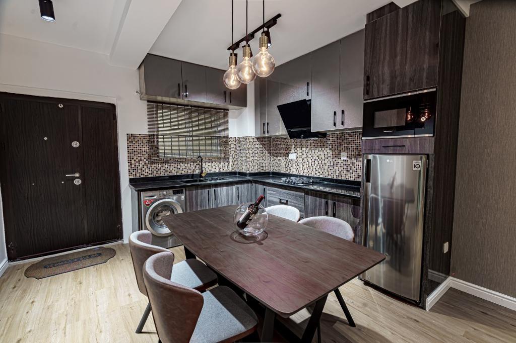 A modern kitchen area showcases sleek cabinetry and a stylish dining table with four chairs. Equipped with contemporary appliances, the kitchen features a washing machine, fridge, and gas hob. Warm lighting from overhead fixtures highlights the textured backsplash and polished countertops.