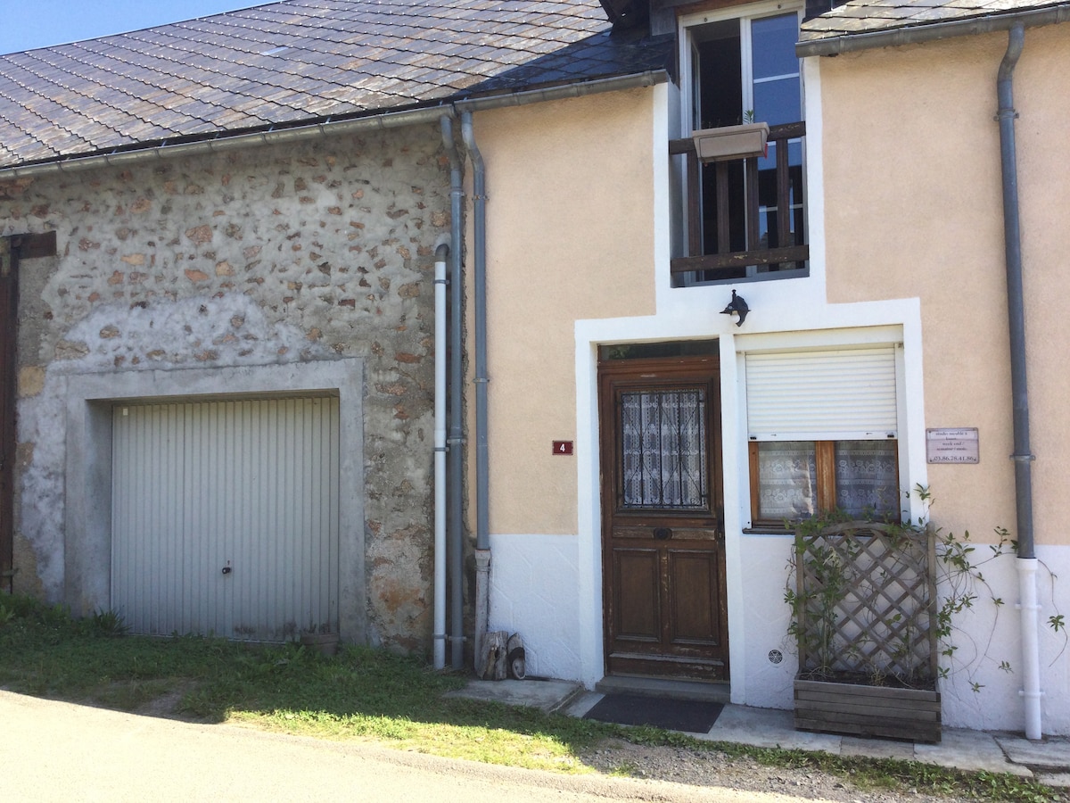 The entrance of a charming house is depicted, featuring a wooden door with a decorative glass panel and a shuttered window above it. The stone and plaster exterior is complemented by a small wooden planter box with greenery. An attached garage is visible to the left.