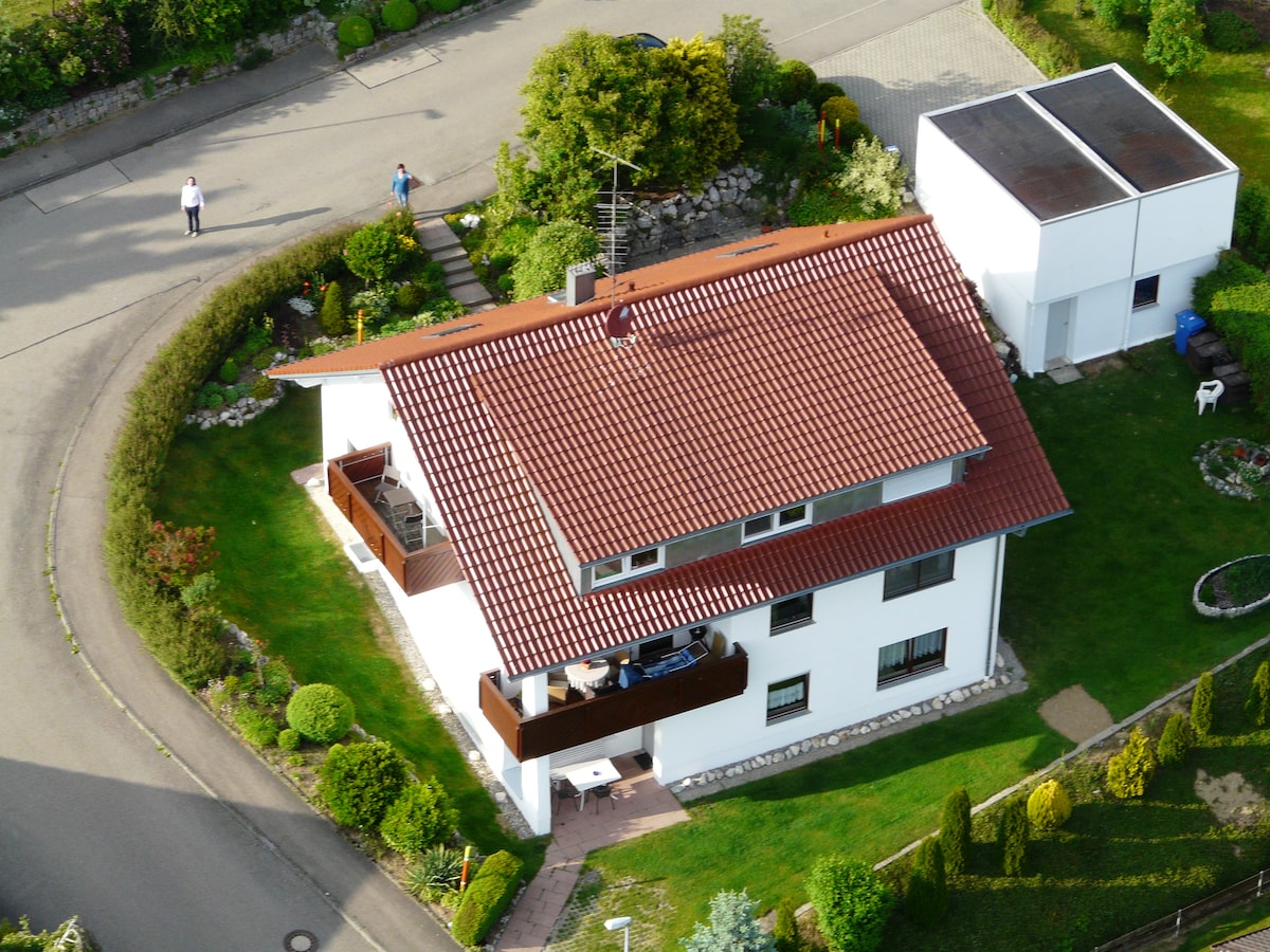 An aerial view showcases a two-story house surrounded by well-maintained greenery. The structure features a red tiled roof and white exterior, with a covered terrace visible on the upper floor. Pathways, shrubs, and neatly trimmed hedges create a serene outdoor space.