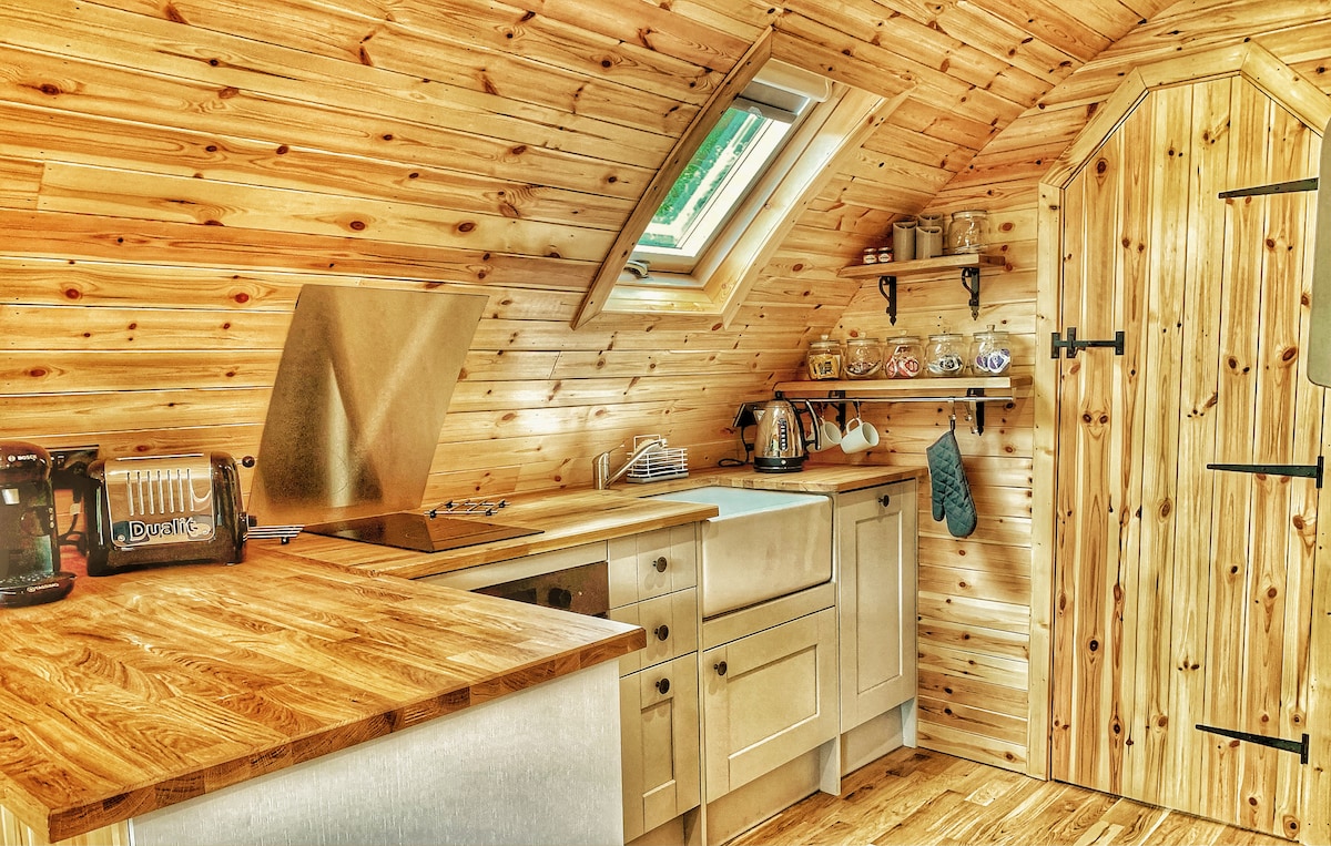 The kitchen features wooden cabinetry and a spacious countertop. Natural light enters through a skylight and a small window. Appliances include a kettle, toaster, and a dishwasher, with shelving showcasing an array of mugs and kitchen essentials.