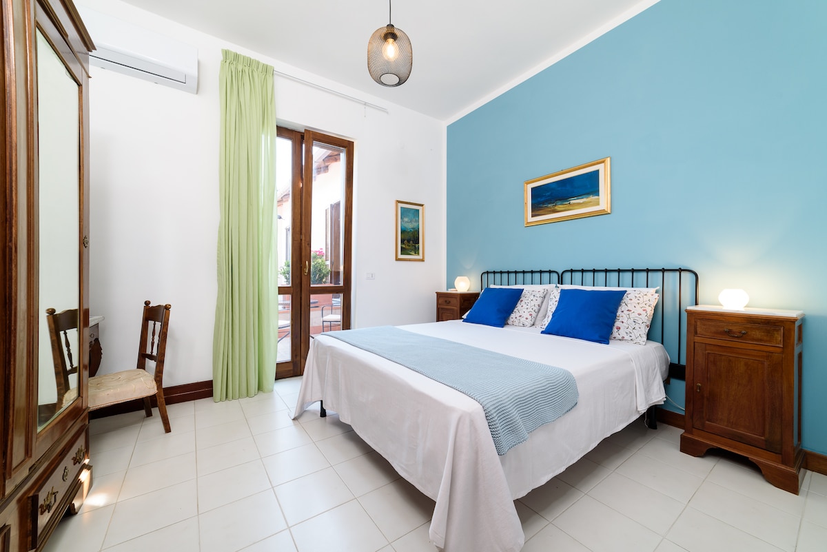 A spacious room features a double bed with a light blue blanket and decorative pillows. A wooden wardrobe and a chair are present. Natural light enters through a door leading to a private terrace, while a soft blue wall complements the decor.
