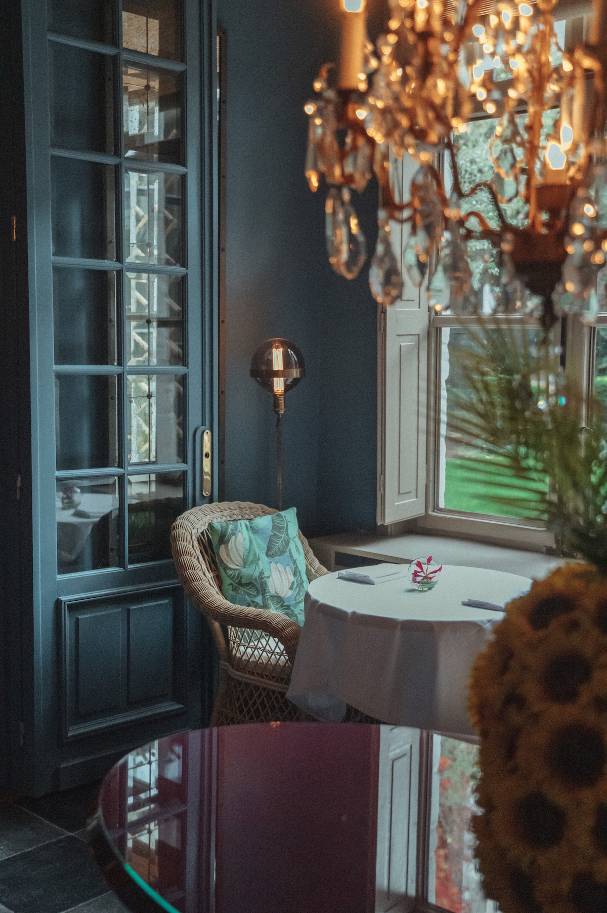 An intimate dining area is featured, with a round table covered in a white tablecloth, surrounded by playful greenery. A large window offers views of the garden, while a chandelier hangs elegantly overhead, casting a warm glow. A wicker chair is adorned with a patterned cushion.