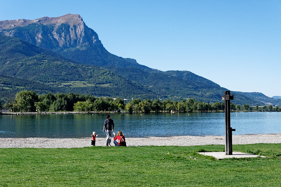 A scenic view of a tranquil lakeside beach with a sandy shore and lush green grass. In the foreground, a family is gathered, enjoying their time by the water's edge. The backdrop features a majestic mountain range under a clear blue sky.