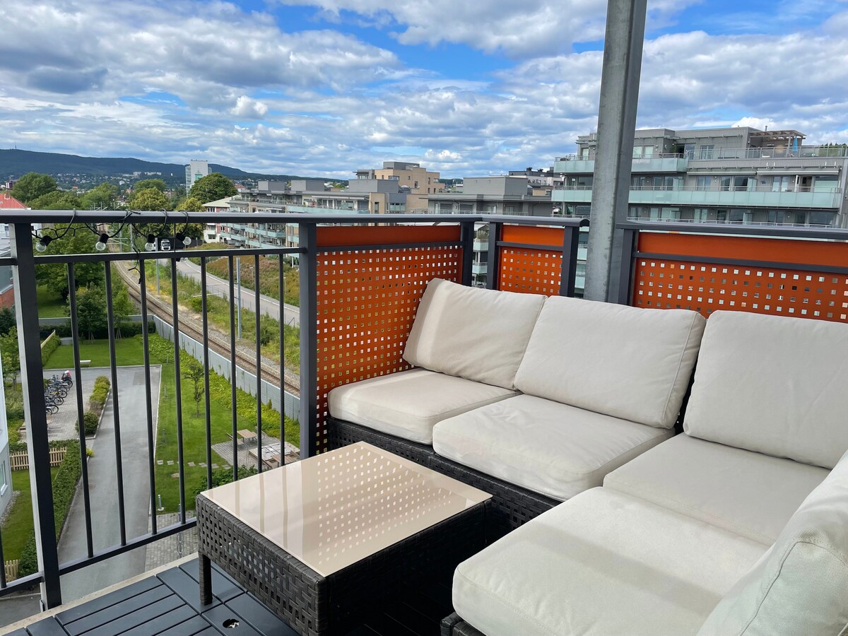 A spacious balcony features a comfortable seating area with light-colored cushions. A sleek table is placed in the center, surrounded by panoramic views of the neighborhood. The lush greenery and distant buildings create a serene backdrop under a partly cloudy sky.