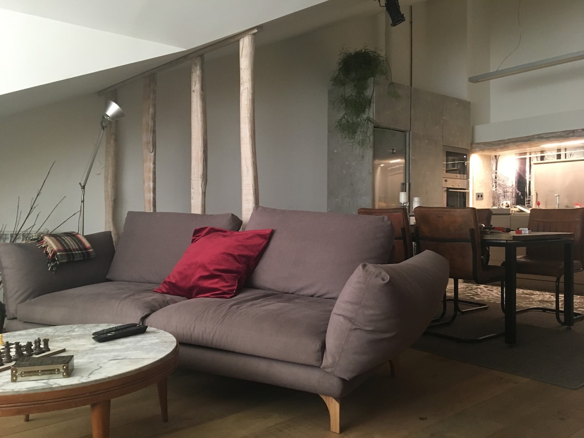 A modern living area is presented, featuring a gray sectional sofa adorned with maroon cushions. A round marble coffee table sits in front, with a chess set displayed. The backdrop includes natural wood accents and a contemporary kitchen visible in the distance.