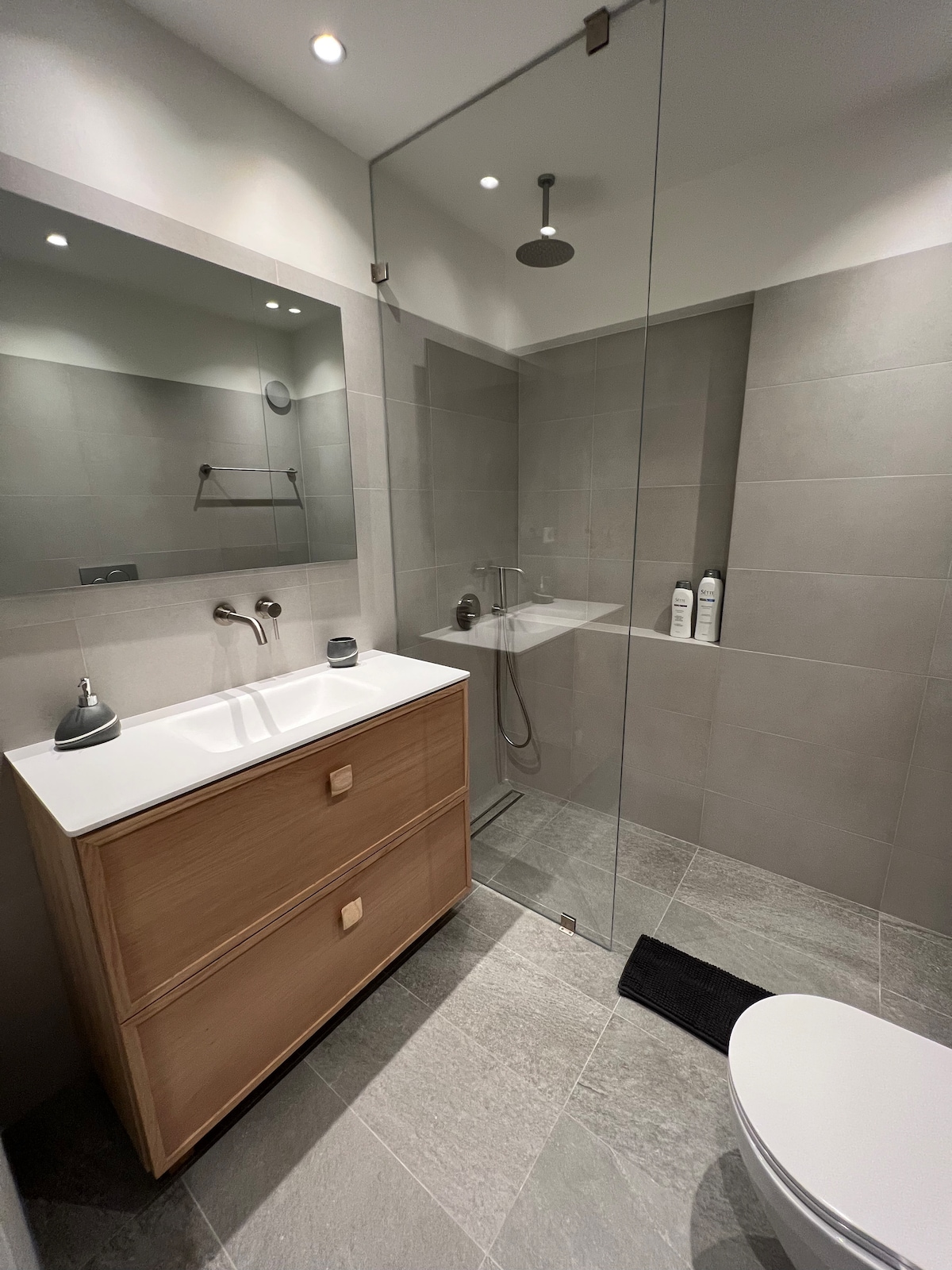 A modern bathroom features a walk-in glass shower with a rain showerhead. A light wood vanity provides dual sinks, complemented by simple fixtures. Neutral grey tiles cover the floor and walls, creating a clean and spacious appearance. A black mat lies in front of the toilet.