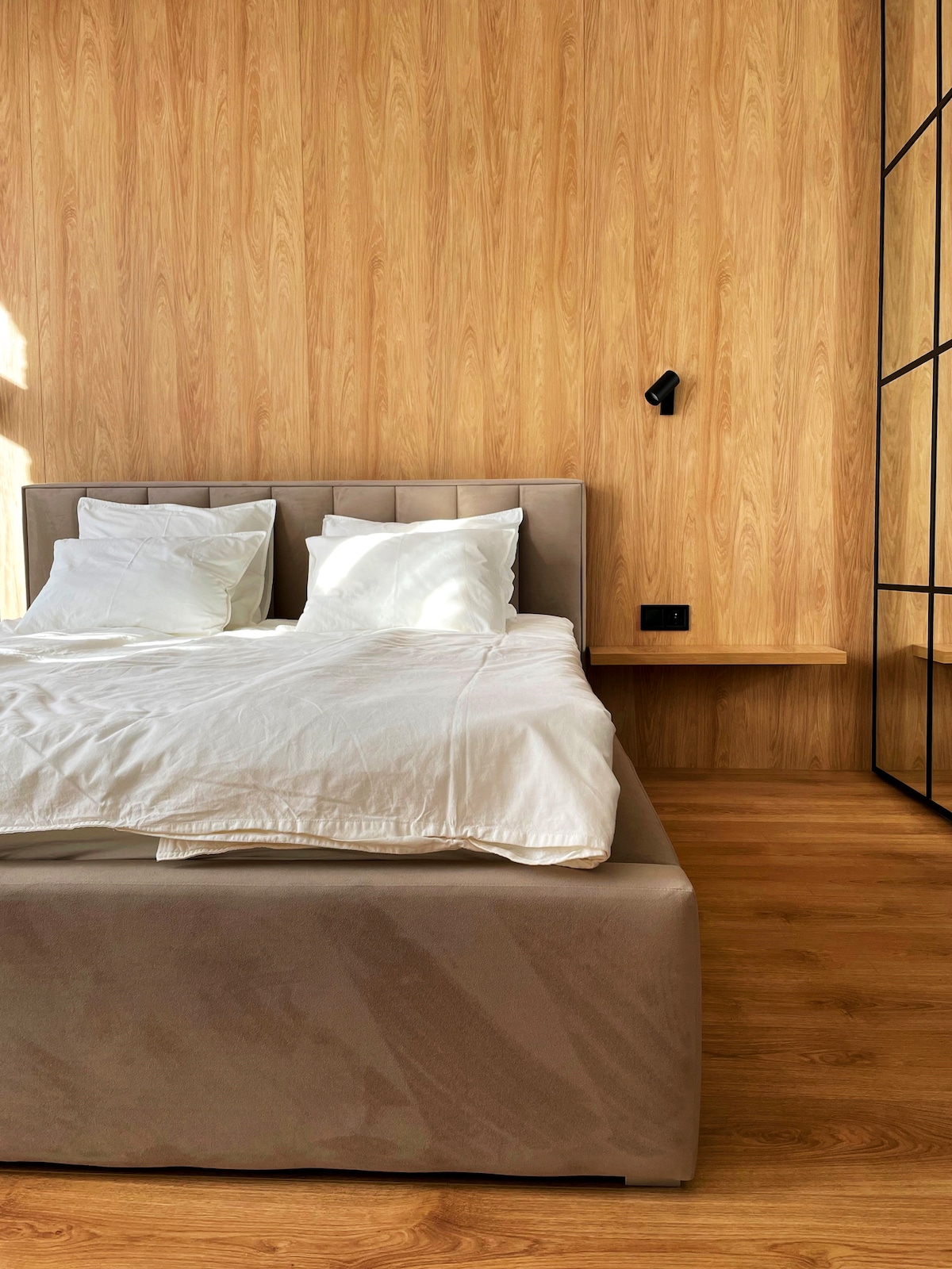 A spacious bed is positioned against a warm wooden wall, featuring crisp white linens and two plush pillows. A small bedside shelf is mounted on the wall beside the bed, while the warm-toned wooden flooring complements the overall tranquil ambiance.