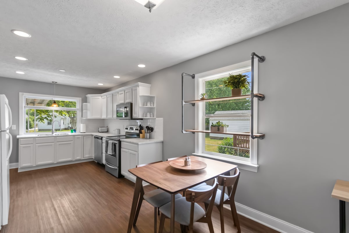 The kitchen features modern stainless steel appliances and white cabinetry, creating a clean and functional space. A small wooden dining table is surrounded by four chairs, with natural light streaming through two windows. Open shelving displays potted plants, adding a touch of greenery.