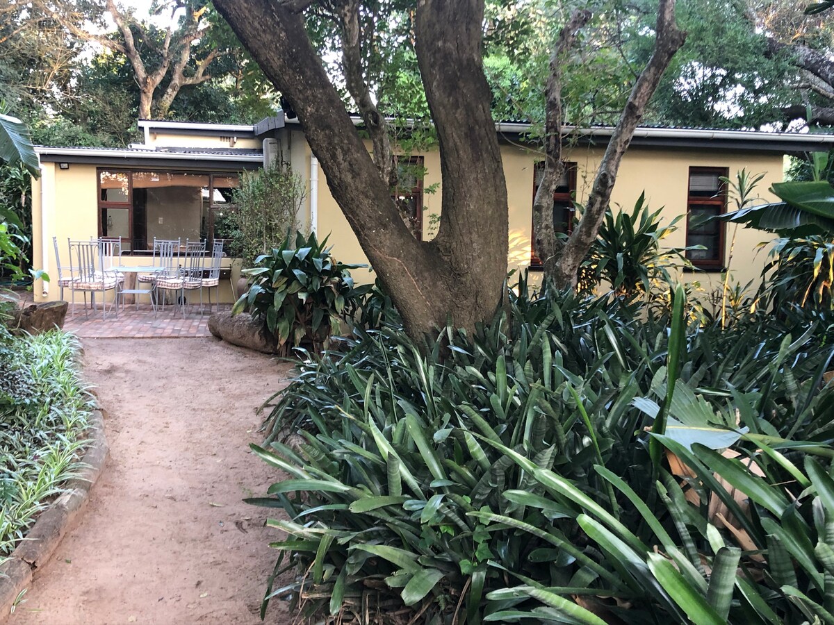 The entrance path leads toward a cottage surrounded by lush greenery and vibrant plants. A spacious outdoor seating area is visible, situated under a tree, with a table and several chairs designed for relaxation. The exterior features soft yellow walls and large windows.