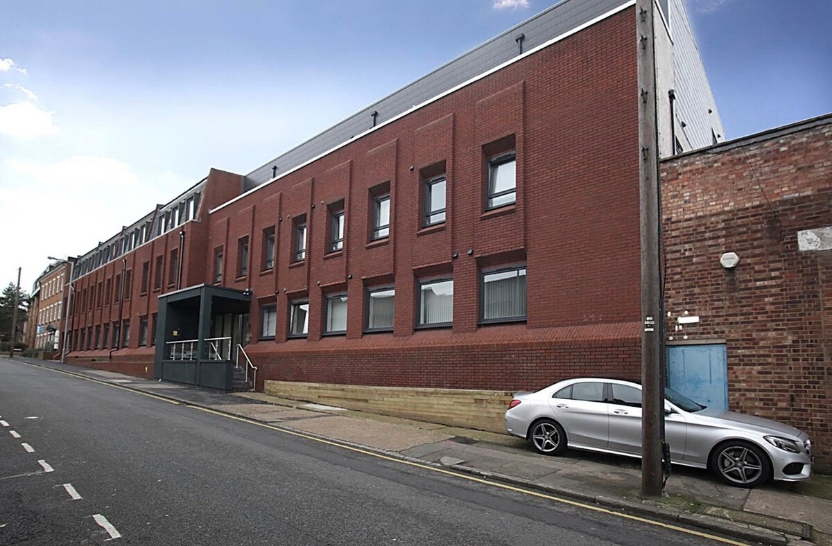 A modern red-brick building is shown from a street view, featuring multiple windows and a prominent entrance with stairs leading up. A parked silver sedan is visible along the side of the building, with an adjacent brick wall adding texture to the scene.