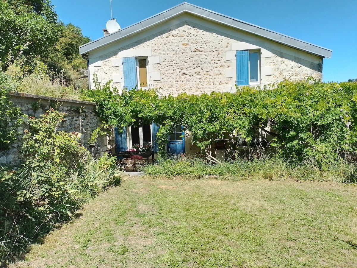 The image shows a charming stone house with blue window shutters surrounded by green vegetation. A well-maintained garden stretches in the foreground, while a terrace and outdoor seating area are visible through the lush foliage, providing a serene outdoor space.