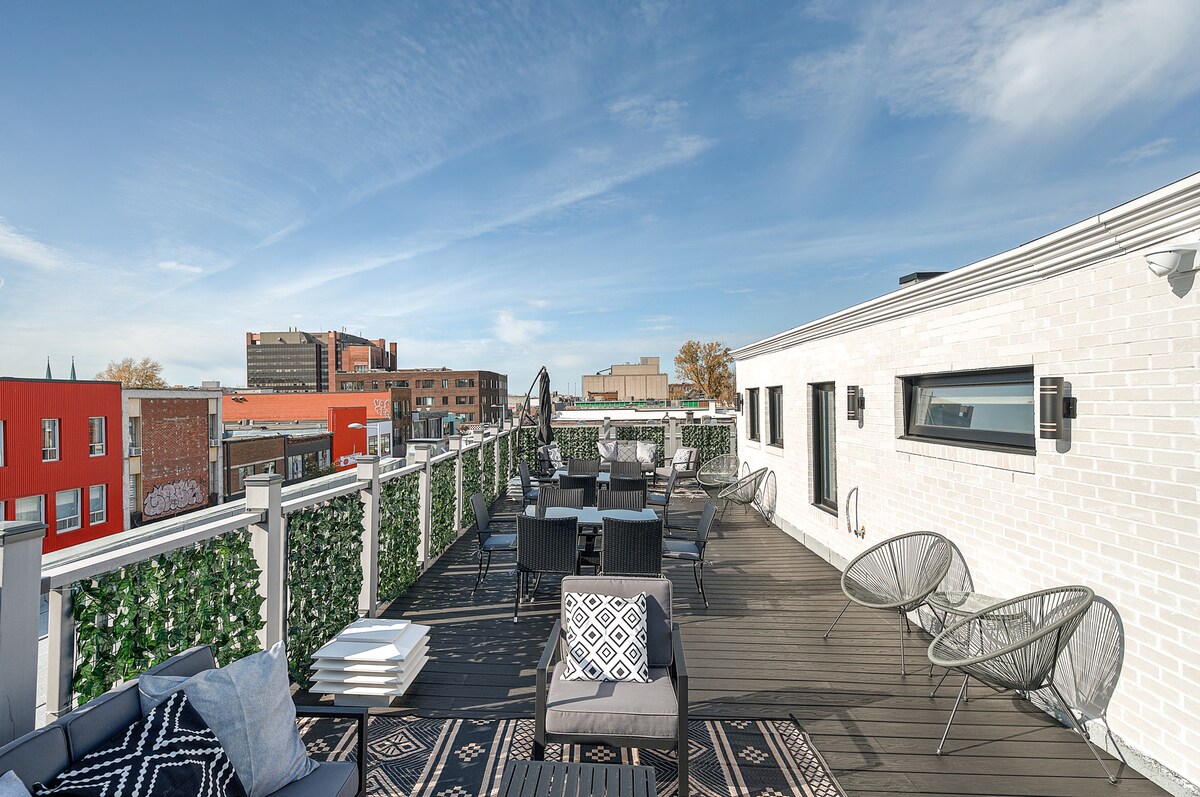 The rooftop terrace features a wooden deck adorned with lounge chairs and tables, providing ample seating for gatherings. Lush greenery borders the space, enhancing privacy. A clear sky stretches above, while city buildings are visible in the background, creating a serene environment for relaxation.