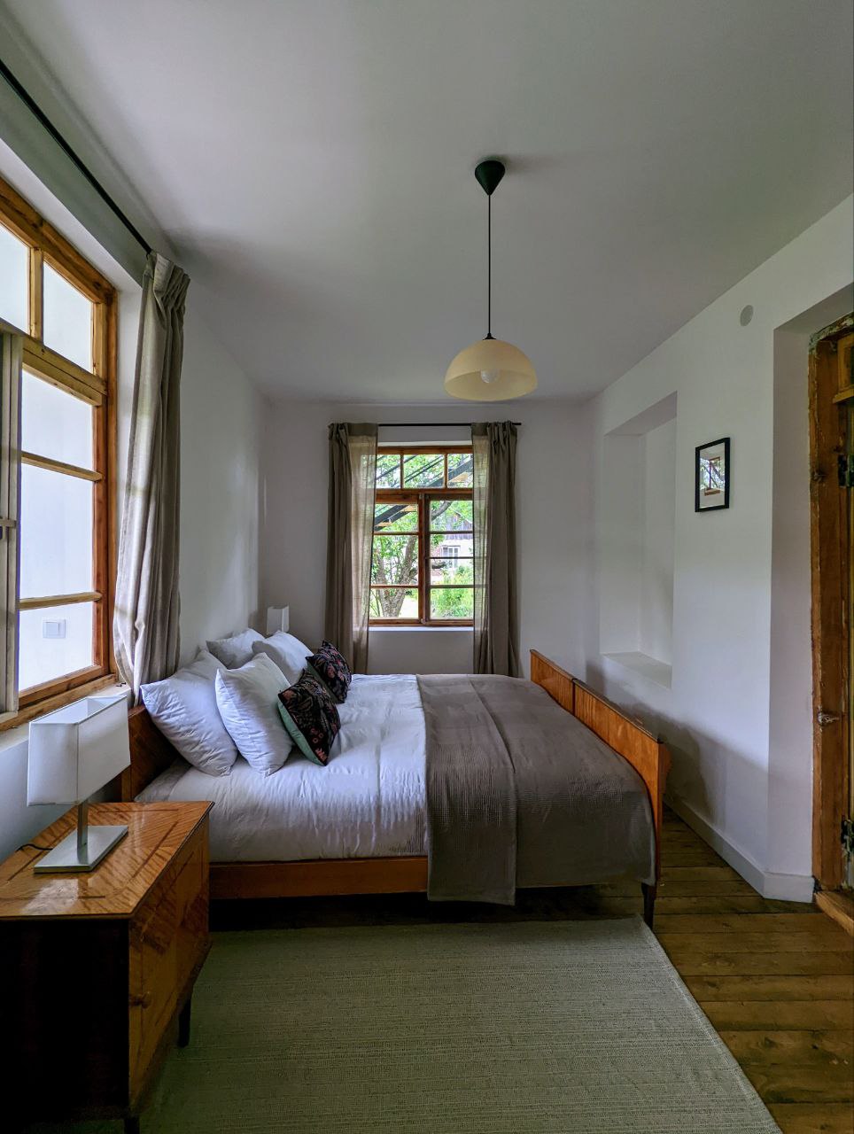 A light-filled bedroom features a spacious bed with crisp white linens, positioned against a window with multiple panes that brings in natural light. A wooden bedside table is placed beside the bed, complementing the warm tones of the room's decor. A neutral area rug covers the wooden floor.