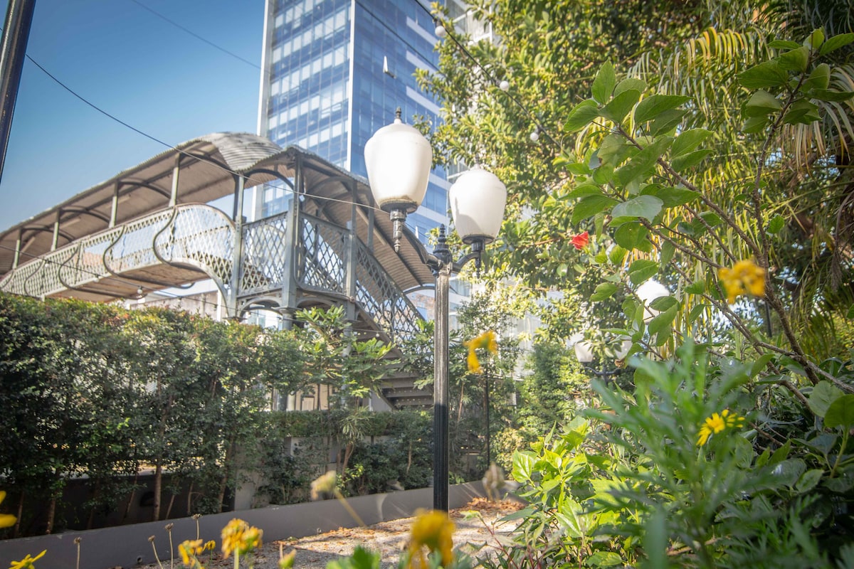 A landscaped courtyard features vibrant greenery and blooming flowers, with ornamental street lamps illuminating the area. A modern building rises in the background, creating a contrast with the natural elements present in the inviting outdoor space.