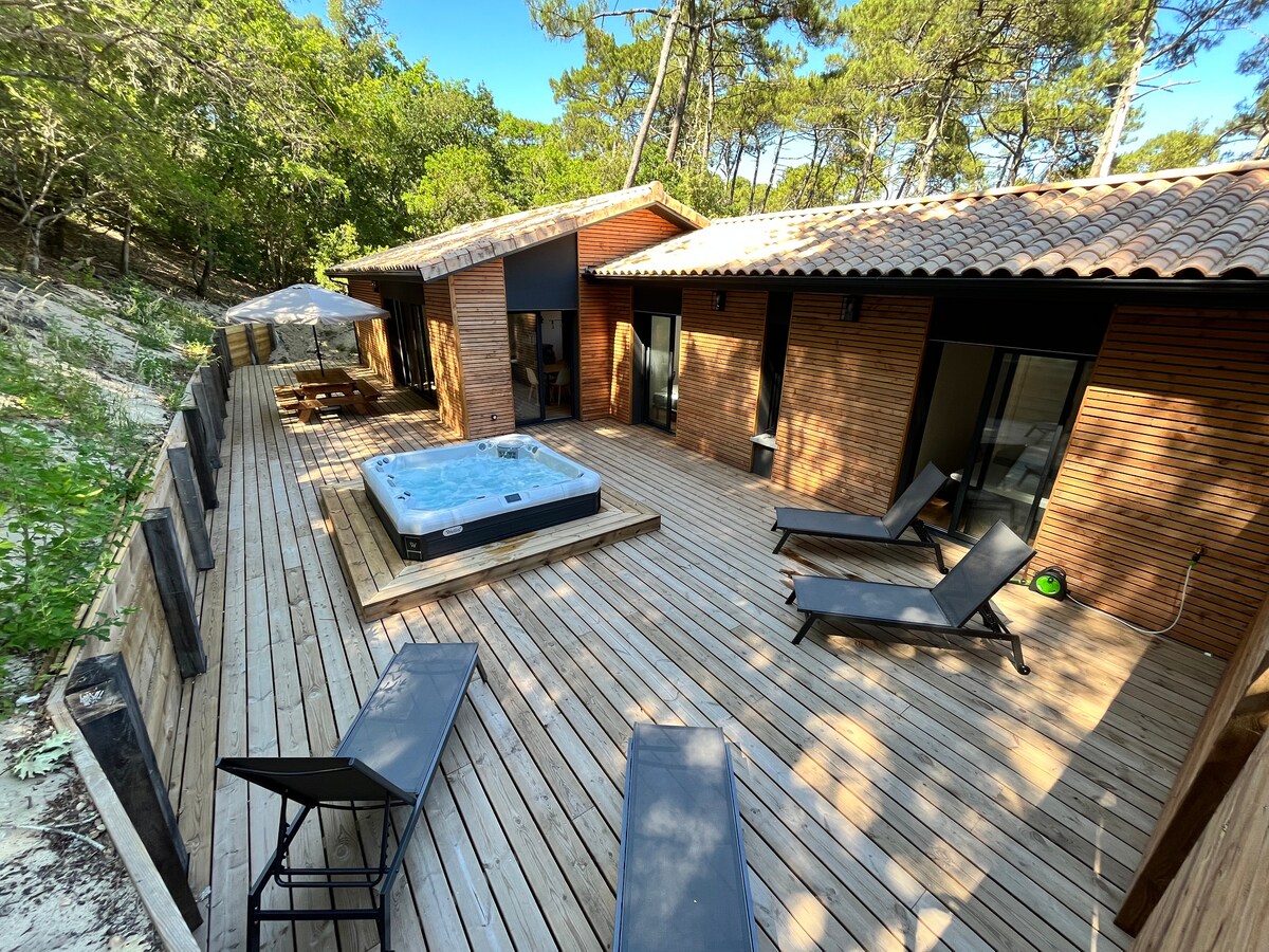 A spacious wooden terrace is enveloped by trees, featuring a hot tub at its center. Lounge chairs are arranged for relaxation, while an umbrella provides shade. Large sliding glass doors lead to the indoor living areas, designed for a seamless connection with the outdoor space.