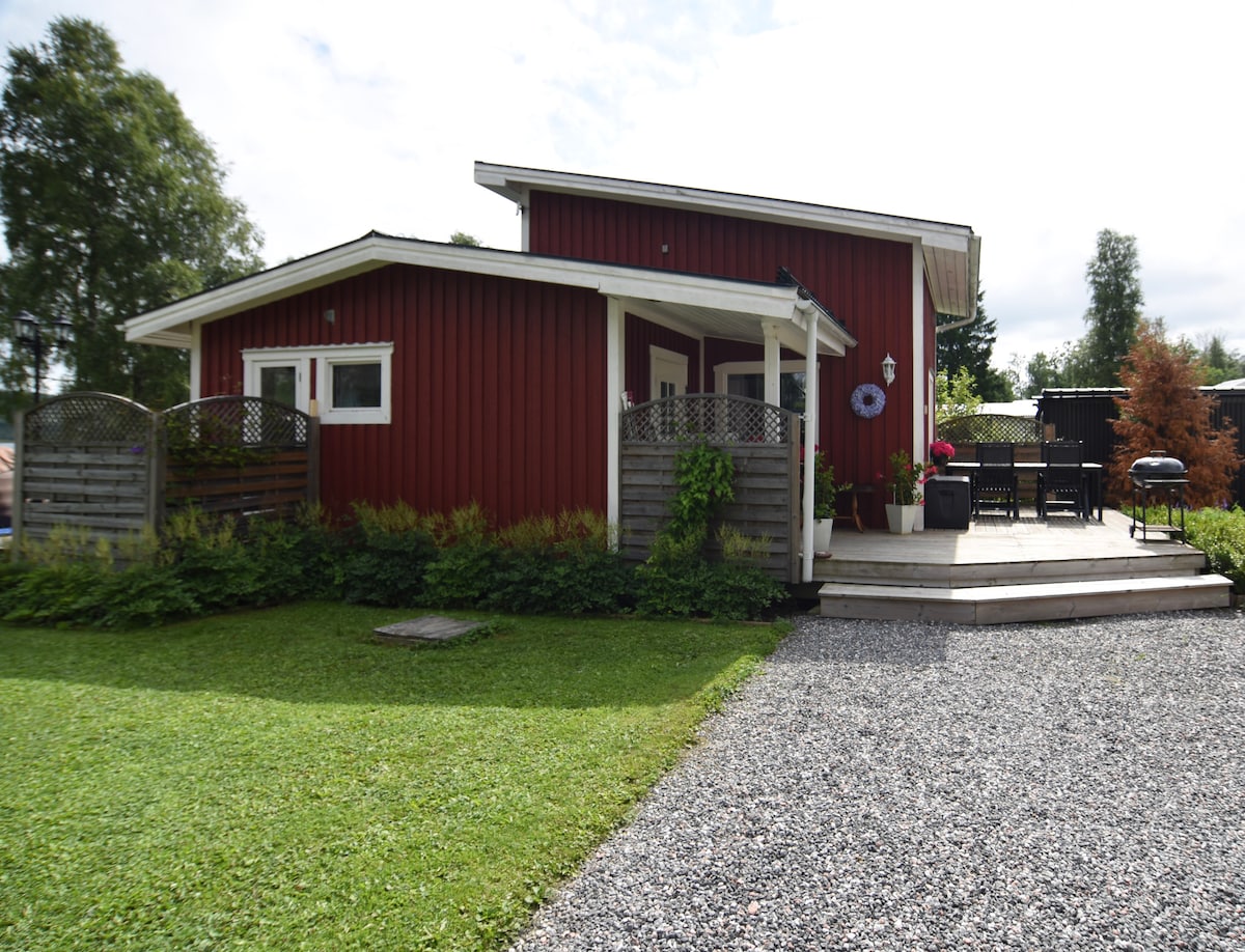 A red wooden house is set against a green lawn, with neatly arranged gravel pathways leading to a welcoming porch. Lush plants frame the entrance, while a patio with outdoor seating is visible beside the house, providing an inviting outdoor space.