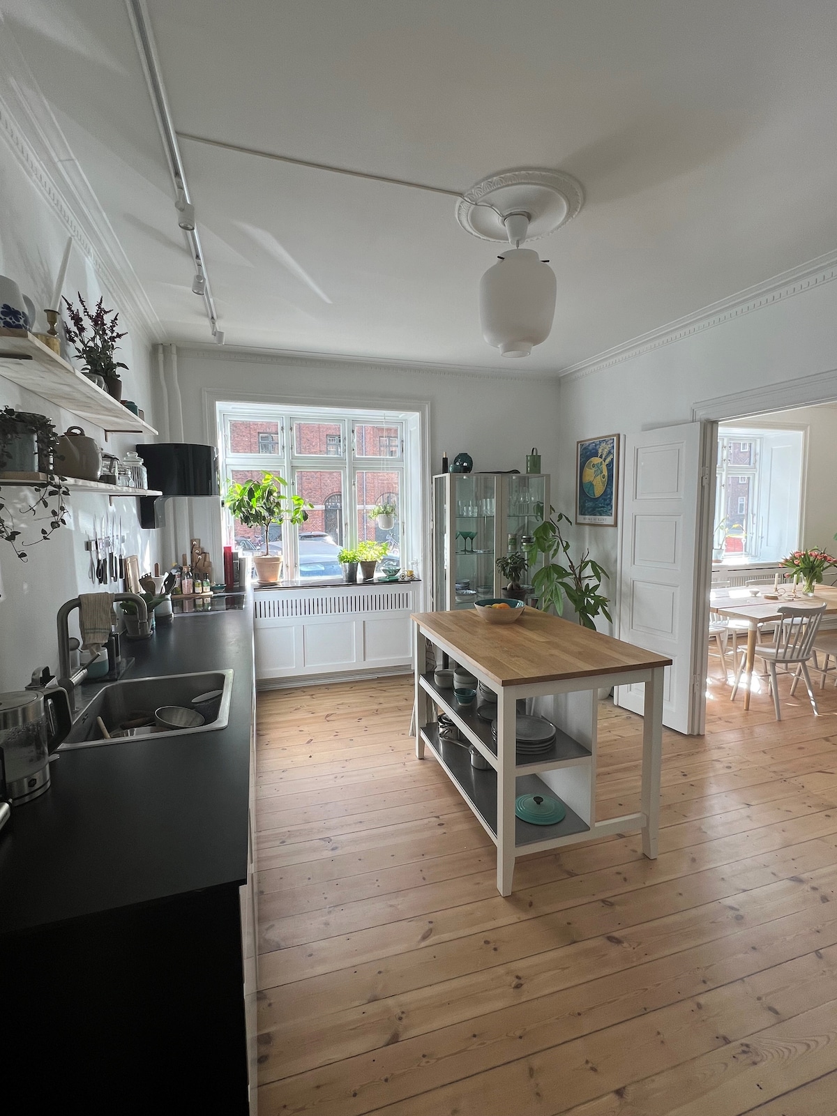 A spacious kitchen includes a central wooden island with seating. Dark countertops contrast with white cabinetry. Natural light floods in through a large window, illuminating potted plants on the windowsill. A glass cabinet displays kitchenware, while a dining area is visible in the background.