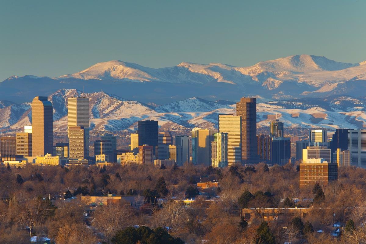 A panoramic view of a city skyline is presented, backed by snow-capped mountains. The buildings vary in height and design, with a mix of glass and concrete structures. A soft golden light highlights the scene, creating a serene contrast against the natural landscape.