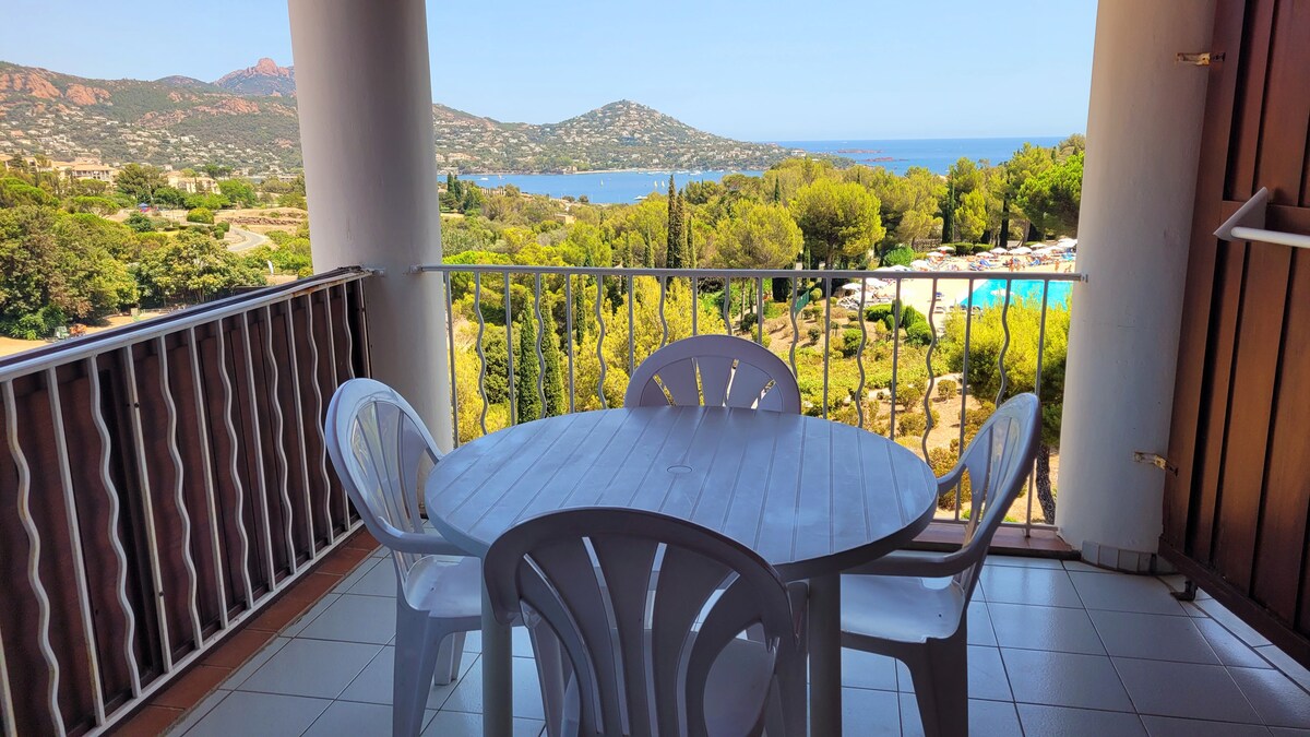 A terrace is visible, featuring a round table surrounded by four white chairs. The space offers a view of lush greenery, swimming pools, and the sea in the distance, framed by scenic hills and a clear blue sky.