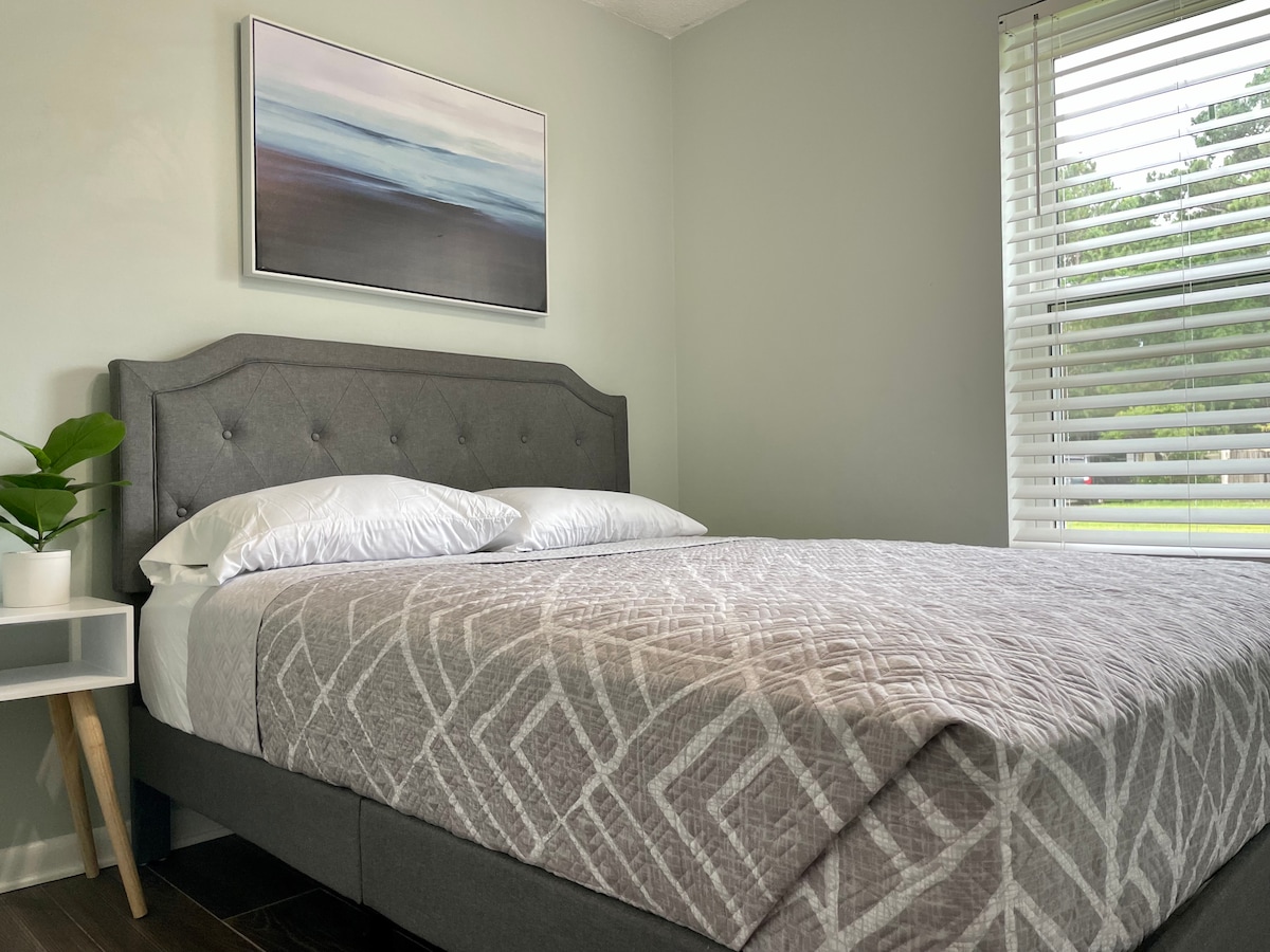 A cozy bedroom features a spacious bed with a gray upholstered headboard and a light-colored quilt. Natural light enters through a window adorned with blinds, illuminating a small nightstand that holds a potted plant. A calming beach-themed artwork hangs above the bed.