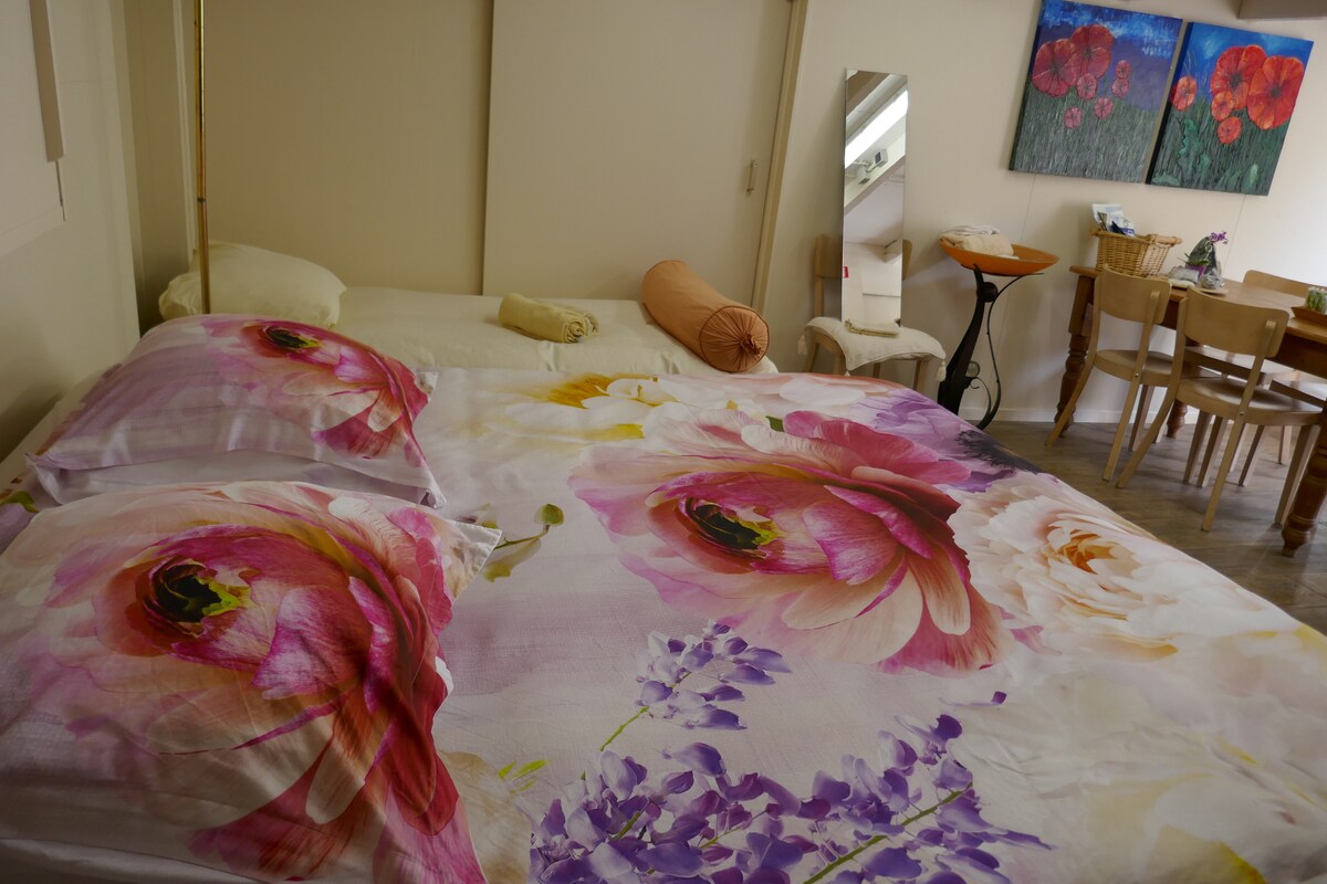 A spacious sleeping area features a large bed adorned with floral-patterned bedding. A mirror is positioned nearby, reflecting the soft colors of the room. To the side, a dining table and chairs complement the cozy atmosphere, with artworks enhancing the visual appeal.