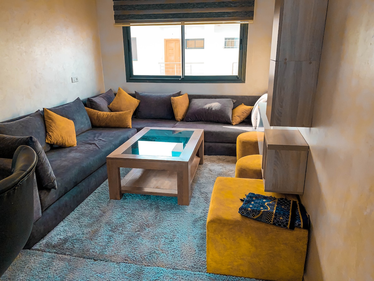 A cozy living area features a dark sectional sofa adorned with yellow accent pillows. A glass-top coffee table is centered on a plush rug. Light enters through a large window, framed by modern shades, brightening the neutral-toned walls.