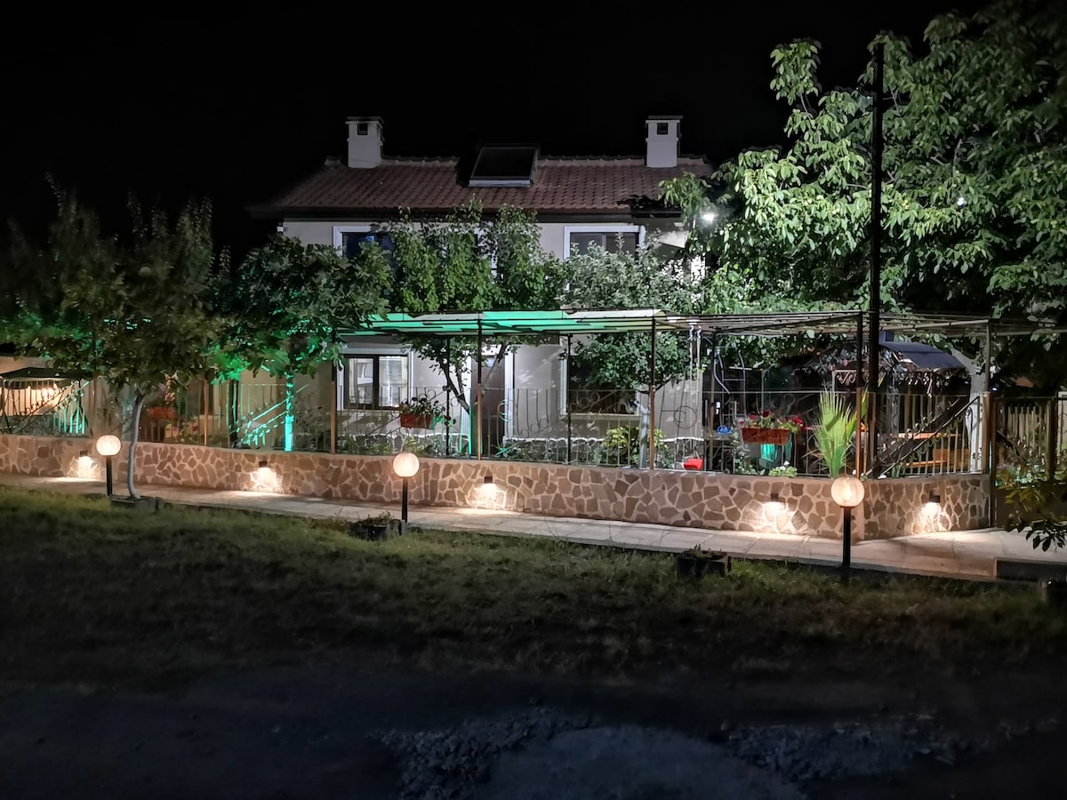 The exterior of the guest house is illuminated at night, highlighting the stone wall and landscaped garden. Lanterns cast a warm glow along the pathway. The house features a covered terrace adorned with plants, providing a cozy atmosphere under the night sky.