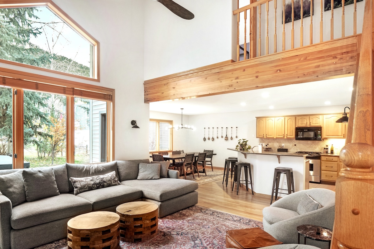 An airy living space features a large sectional sofa arranged around rustic wooden coffee tables. A view of the dining area and kitchen can be seen in the background, with modern bar stools positioned near the counter. Natural light floods through expansive windows.