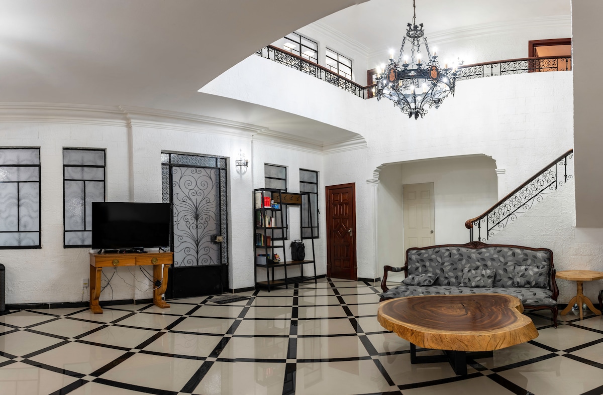 An elegant living area features polished tile flooring with a geometric pattern. A vintage chandelier hangs overhead, illuminating the space. A large wooden table sits at the center, surrounded by a gray sofa. An intricate iron railing leads to the upper level.