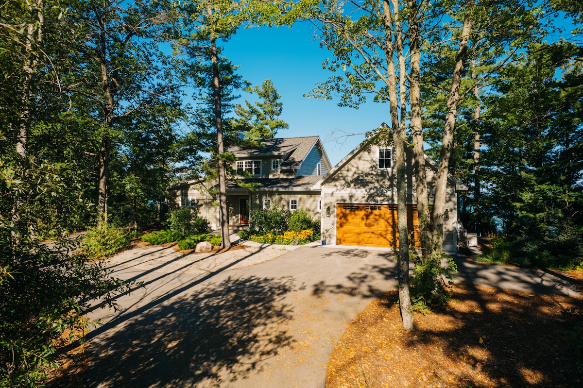 The exterior of a modern home is framed by trees and greenery, showcasing a blend of natural surroundings and architecture. A driveway leads to a wooden garage door, while a clear blue sky provides a bright backdrop, enhancing the overall serene ambiance.
